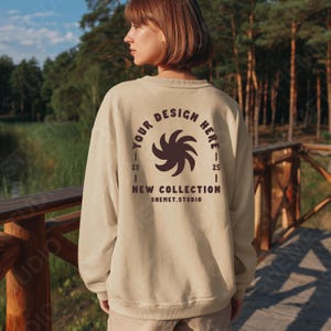 May include: Beige sweatshirt with a brown graphic design on the back. The design includes the text "YOUR DESIGN HERE", "NEW COLLECTION", and "SHEMET.STUDIO". The sweatshirt is worn by a person standing on a wooden bridge.