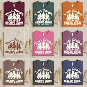 Op de afbeelding: Negen verschillend gekleurde t-shirts worden getoond, elk netjes opgevouwen. De shirts hebben een ronde hals en de tekst "Your Design" boven een afbeelding van dennenbomen en de woorden "MOCKUPS STUDIO High-Quality Mockups."