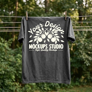 以下が含まれることがあります： 洗濯ロープに吊るされた濃い灰色のTシャツ。Tシャツには、果物のある枝のデザインの上に「Your Design」というテキストと、「MOCKUPS STUDIO - High-Quality Mockups」というテキストが白で描かれています。