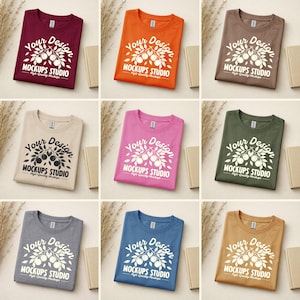 May include: Nine folded t-shirts in various colors, including burgundy, orange, and brown, are displayed. Each shirt features the text "Your Design" and "MOCKUPS STUDIO" in white, along with a graphic of a branch with fruit. The shirts are arranged on a neutral surface.