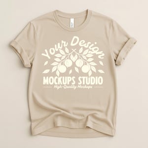Könnte beinhalten: Beiges T-Shirt mit hochgekrempelten Ärmeln. Das Shirt zeigt den Text "Your Design" über einer Grafik aus Blättern und Früchten, darunter "MOCKUPS STUDIO" und "High-Quality Mockups". Das T-Shirt ist aus weichem Material.
