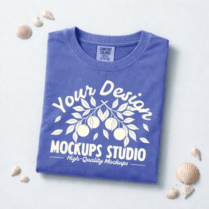 May include: A folded blue t-shirt with the text "Your Design MOCKUPS STUDIO High-Quality Mockups" in white. The design includes a branch with leaves and fruit. The shirt is surrounded by seashells.