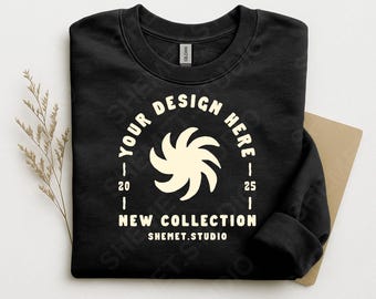 May include: Black crewneck sweatshirt with the text "YOUR DESIGN HERE" in a circular design, with a cream-colored graphic in the center. The text "NEW COLLECTION" and "SHEMET.STUDIO" are also visible. The sweatshirt is folded.