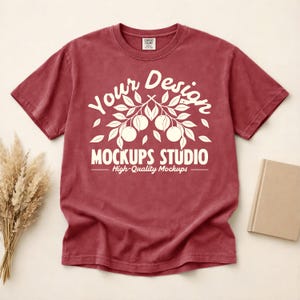 Könnte beinhalten: Ein weinrotes T-Shirt mit Rundhalsausschnitt und kurzen Ärmeln. Das Shirt hat eine weiße Grafik mit dem Text "Your Design MOCKUPS STUDIO High-Quality Mockups". Das Shirt wird vor einem neutralen Hintergrund präsentiert.