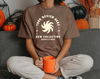 May include: A brown t-shirt with the text "YOUR DESIGN HERE" and "NEW COLLECTION SHEMET.STUDIO" in white. An orange mug is held in the hands. The person is wearing light-colored pants and black and white checkered shoes. Pumpkins and fall decor are in the background.