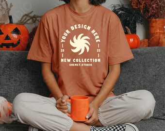 May include: A rust-colored t-shirt with the text "YOUR DESIGN HERE" and "NEW COLLECTION" in a white font. The person is holding an orange mug. The background includes a carved pumpkin and fall decorations.