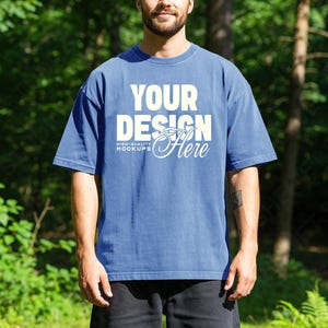 Può includere: Una maglietta blu con la scritta "YOUR DESIGN Here" in bianco. La maglietta ha una vestibilità ampia e maniche corte. Le parole "HIGH-QUALITY MOCKUPS" sono sotto. Il modello indossa pantaloncini neri.