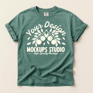 Puede incluir: Una camiseta de color verde azulado con las mangas enrolladas. La camiseta presenta un diseño gráfico blanco con las palabras "Your Design" encima de "MOCKUPS STUDIO" y "High-Quality Mockups" debajo. La camiseta es de material suave.
