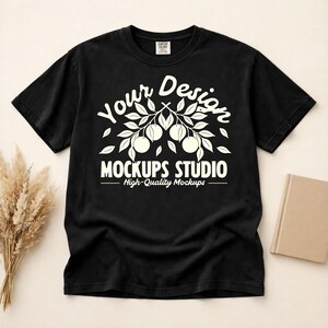 May include: Black t-shirt with a white graphic design. The design features the text "Your Design" above a branch with fruit and the text "Mockups Studio - High-Quality Mockups" below. The t-shirt is on a neutral background.