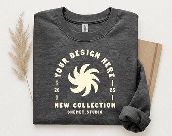 May include: Dark gray sweatshirt with the text "YOUR DESIGN HERE" in a circular design, with a stylized swirl graphic. The text "NEW COLLECTION" and "SHEMET.STUDIO" are also visible. The sweatshirt is folded, with a neutral background.