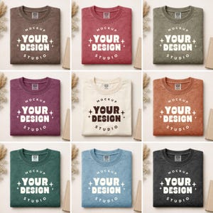 May include: Nine folded t-shirts in various colors, including brown, red, green, purple, cream, and blue. Each shirt features the text "MOCKUP YOUR DESIGN STUDIO" in a white, stylized font. The shirts are arranged on a neutral background.