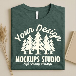 Puede incluir: Camiseta verde oscuro doblada con un gráfico blanco de pinos y el texto "Your Design MOCKUPS STUDIO High-Quality Mockups". La camiseta tiene una etiqueta en el cuello.