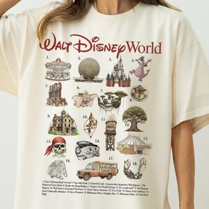 Puede incluir: Camiseta extragrande de color crema con un gr&aacute;fico de las atracciones de Walt Disney World. La camiseta incluye el texto "Walt Disney World" en rojo, con ilustraciones numeradas de varios puntos de referencia del parque.