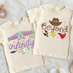 Puede incluir: Dos camisetas crema de manga corta con dise&ntilde;os gr&aacute;ficos. Una camiseta presenta una nave espacial morada y verde con el texto "To infinity". La otra camiseta tiene un sombrero de vaquero marr&oacute;n y el texto "and Beyond".
