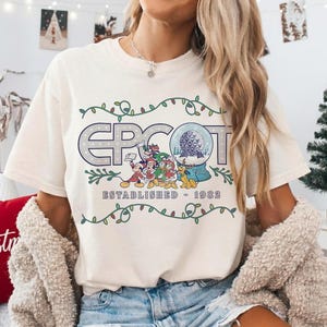 May include: Cream-colored t-shirt featuring the word "EPCOT" in a stylized font, with a snow globe graphic and Disney characters. The text "ESTABLISHED - 1982" is below. The design is framed by a green vine with white berries.