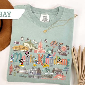 Op de afbeelding: Lichtgroen T-shirt met een kleurrijke afbeelding van het Magic Kingdom met Disney-figuren. De tekst "Magic Kingdom" is weergegeven in een speels lettertype. Een gouden ketting ligt over het shirt.