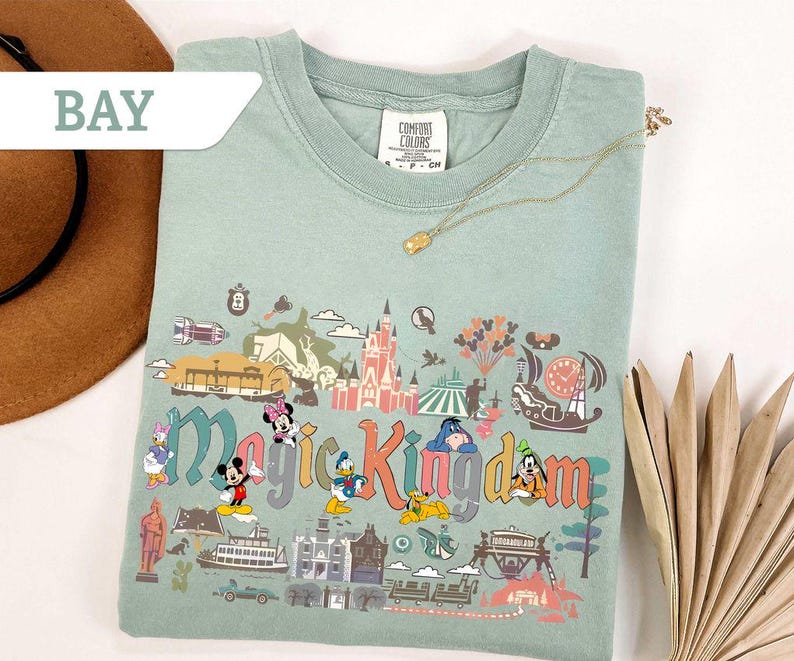 Op de afbeelding: Lichtgroen T-shirt met een kleurrijke afbeelding van het Magic Kingdom met Disney-figuren. De tekst "Magic Kingdom" is weergegeven in een speels lettertype. Een gouden ketting ligt over het shirt.
