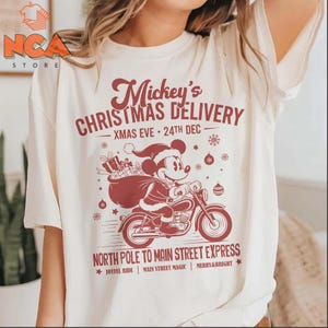 May include: A cream-coloured t-shirt with a red graphic of Mickey Mouse on a motorbike, carrying a sack of presents. The text says "Mickey's Christmas Delivery - Xmas Eve - 24th Dec" and "North Pole to Main Street Express".