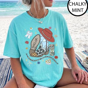 May include: A chalky mint short-sleeve t-shirt featuring a graphic print of a cowboy boot with a cow print pattern, a red hat, blocks with the letters T and S, and the name Taylor. The design includes small flowers, bows, and a beaded string.