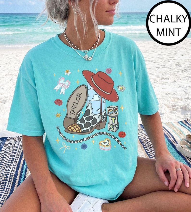 May include: A chalky mint short-sleeve t-shirt featuring a graphic print of a cowboy boot with a cow print pattern, a red hat, blocks with the letters T and S, and the name Taylor. The design includes small flowers, bows, and a beaded string.
