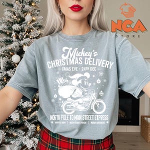 May include: A pale blue t-shirt with a white graphic of Mickey Mouse on a motorbike, delivering presents. The text says "Mickey's Christmas Delivery" and "North Pole to Main Street Express". A festive design for the holiday season.