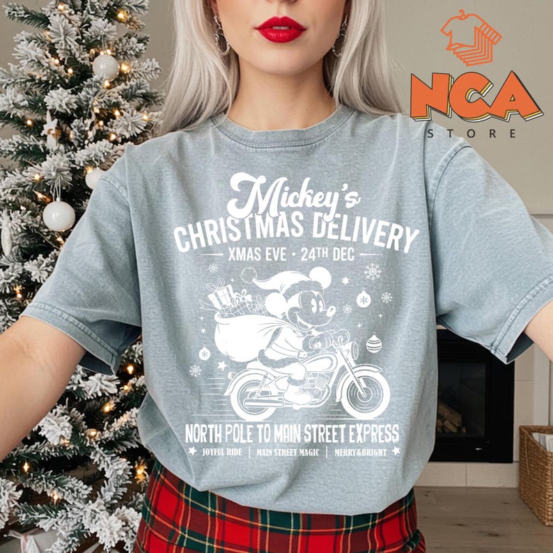 May include: A pale blue t-shirt with a white graphic of Mickey Mouse on a motorbike, delivering presents. The text says "Mickey's Christmas Delivery" and "North Pole to Main Street Express". A festive design for the holiday season.
