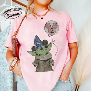 May include: Pink t-shirt with a graphic of a green alien-like character wearing a wizard hat and holding a balloon with the number 2. The shirt has the words "Comfort Colors Blossom" printed on it.