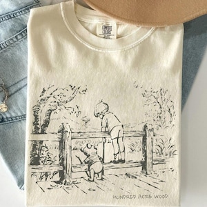 May include: Cream-colored t-shirt featuring a black and white illustration of a boy and a bear looking over a wooden fence. The text "HUNDRED ACRE WOOD" is printed at the bottom of the graphic. The shirt has a crew neck and the brand "COMFORT COLORS" is visible.