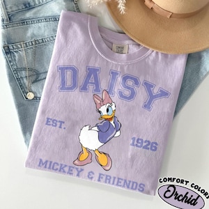May include: A light purple t-shirt featuring a Daisy Duck graphic with the word "DAISY" in large, light purple letters. The shirt also includes the text "EST. 1926" and "MICKEY & FRIENDS". A tan hat and blue jeans are in the background.