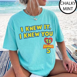 May include: A chalky mint short-sleeve t-shirt featuring the yellow text I KNEW IT, I KNEW YOU above a red heart containing the letters TS, with the word STORY and the number 5 printed in yellow below the heart on a rectangular orange background.