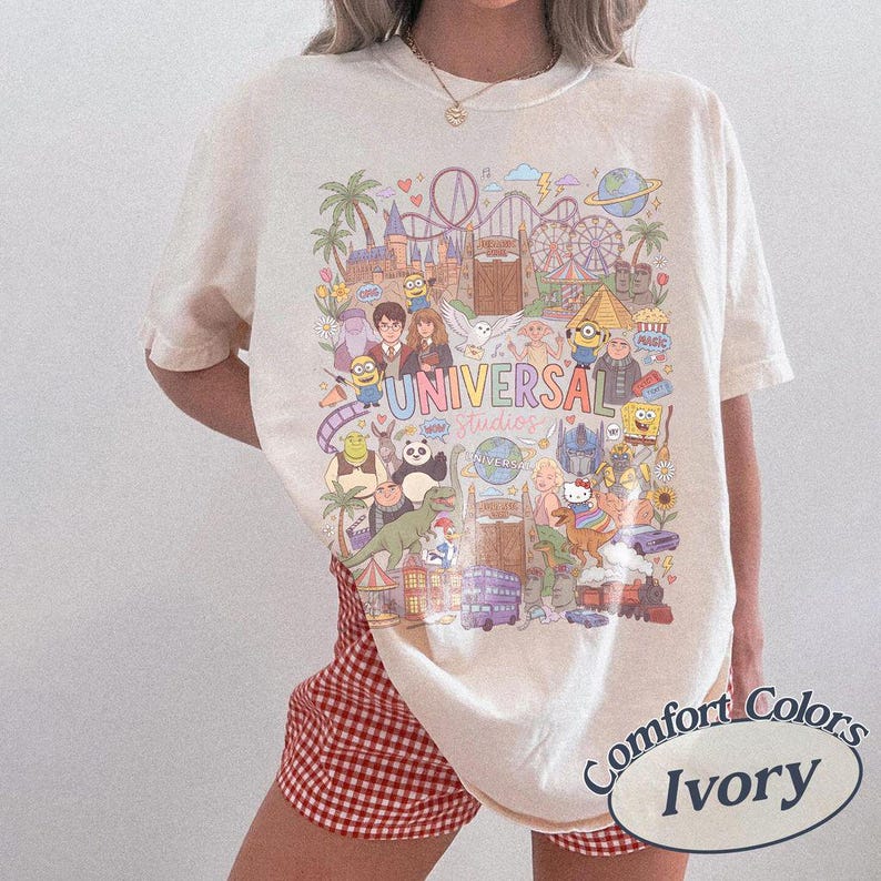 Może przedstawiać: T-shirt z kr&oacute;tkim rękawem w kolorze kości słoniowej z kolorowym nadrukiem graficznym przedstawiającym postacie i punkty orientacyjne Universal Studios. Wz&oacute;r zawiera postacie takie jak Harry Potter, Minionki, Shrek i SpongeBob oraz napis UNIVERSAL Studios.