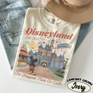 以下が含まれることがあります： アイボリー色のTシャツ。ディズニーランドのグラフィックがプリントされています。デザインには、ディズニーランドのロゴ、お城、花火、ミッキーマウスが描かれています。「Disneyland Est. 1955」と「The Happiest Place On Earth」の文字。