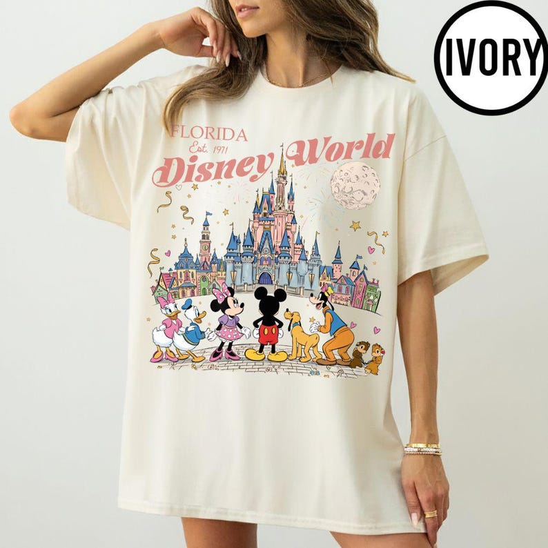 Puede incluir: Camiseta de color marfil con un gr&aacute;fico colorido de Disney World en Florida. El dise&ntilde;o incluye el logotipo de Disney World, un castillo y personajes de dibujos animados como Mickey y Minnie Mouse, Donald Duck y Goofy.