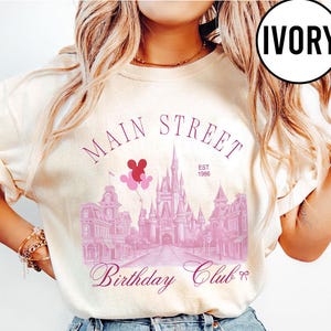 May include: Ivory-colored sweatshirt featuring a Main Street design with a castle, buildings, and balloons. The text includes "Main Street," "Birthday Club," and "EST 1986." The design is in shades of pink.