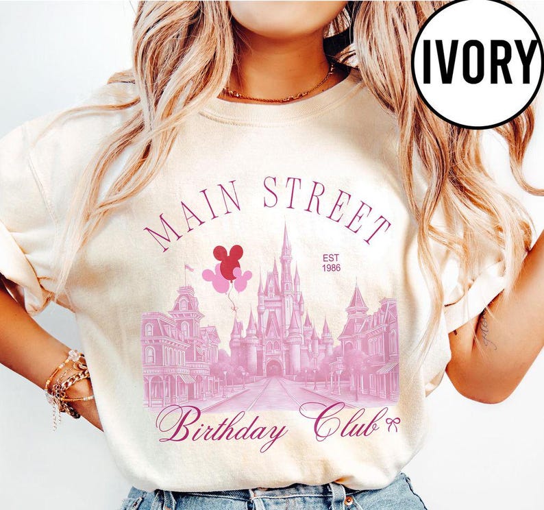May include: Ivory-colored sweatshirt featuring a Main Street design with a castle, buildings, and balloons. The text includes "Main Street," "Birthday Club," and "EST 1986." The design is in shades of pink.