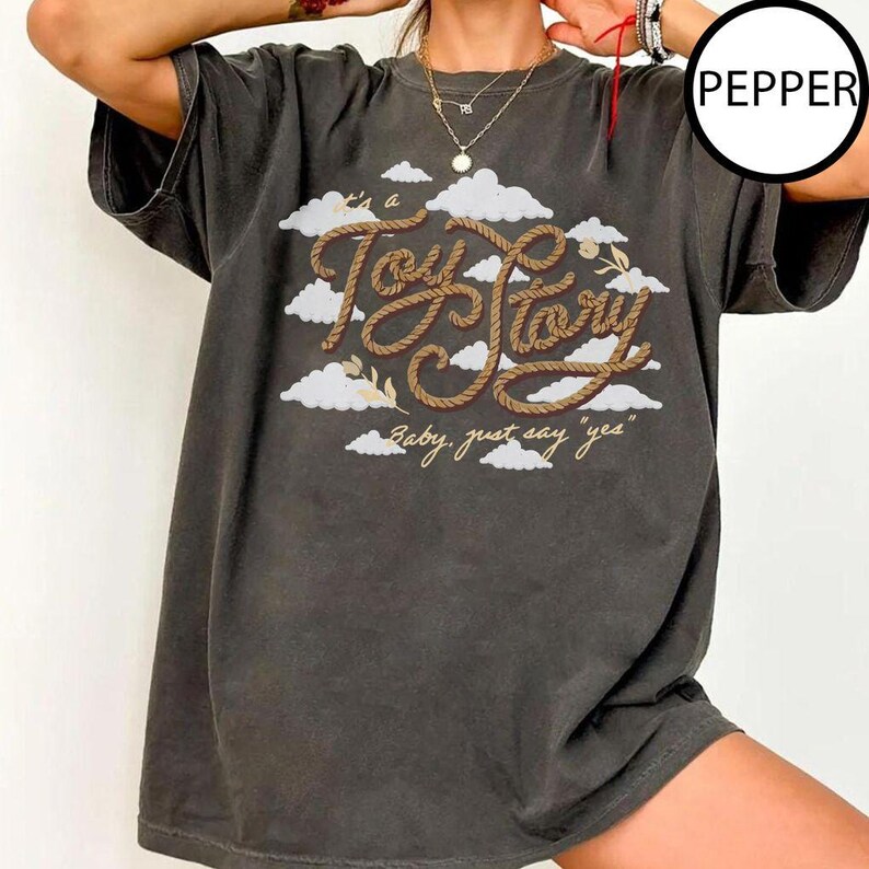 May include: A charcoal gray short-sleeve t-shirt featuring a graphic print of white clouds and the text "It's a Toy Story" written in a brown rope-like font, with the additional text "Baby, just say 'yes'" printed below in a smaller script.