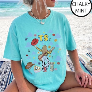 May include: A chalky mint t-shirt featuring a graphic print of a person with long hair playing a guitar, wearing a yellow plaid shirt and cow-print pants. The design includes the text TS, a red hat, a brown cowboy hat, and blue clouds.