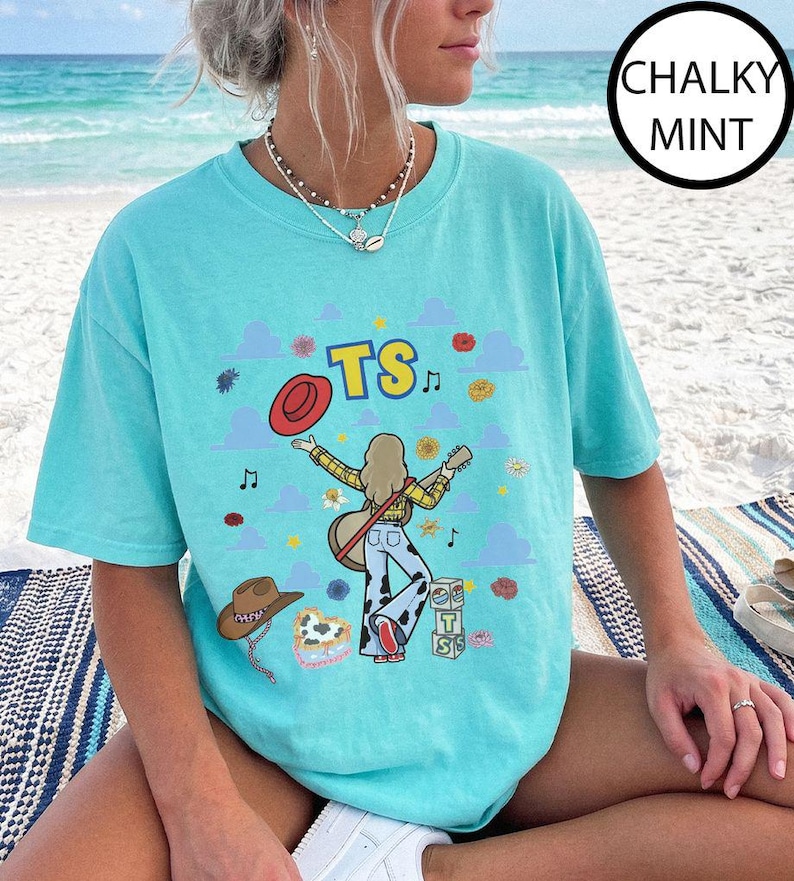 May include: A chalky mint t-shirt featuring a graphic print of a person with long hair playing a guitar, wearing a yellow plaid shirt and cow-print pants. The design includes the text TS, a red hat, a brown cowboy hat, and blue clouds.