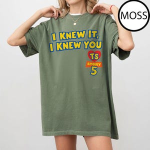 May include: An olive green short-sleeve t-shirt featuring the yellow text I KNEW IT, I KNEW YOU, a red heart with the letters TS, and the orange text STORY 5. The shirt has a crew neckline and a relaxed fit, with a circular MOSS label in the corner.