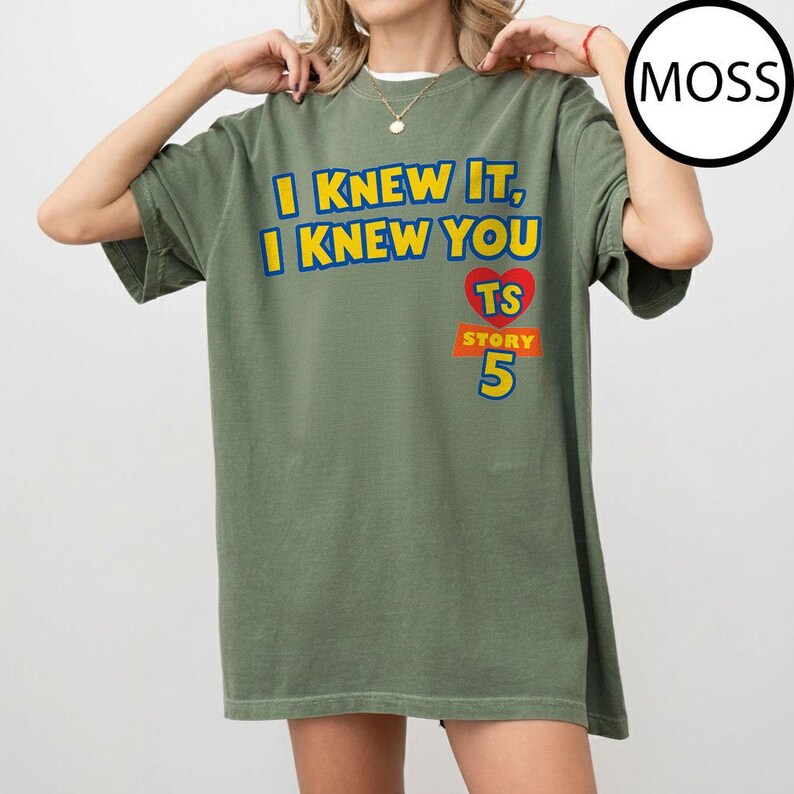 May include: An olive green short-sleeve t-shirt featuring the yellow text I KNEW IT, I KNEW YOU, a red heart with the letters TS, and the orange text STORY 5. The shirt has a crew neckline and a relaxed fit, with a circular MOSS label in the corner.