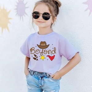 Puede incluir: Una camiseta de color lila claro con la frase "and Beyond" y gr&aacute;ficos tem&aacute;ticos de vaqueros. El dise&ntilde;o incluye un sombrero de vaquero marr&oacute;n, huellas, una estrella amarilla y una bandana roja. La camiseta la lleva un ni&ntilde;o peque&ntilde;o.