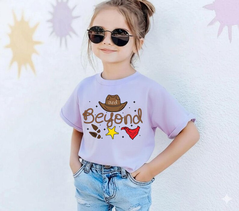 Puede incluir: Una camiseta de color lila claro con la frase "and Beyond" y gr&aacute;ficos tem&aacute;ticos de vaqueros. El dise&ntilde;o incluye un sombrero de vaquero marr&oacute;n, huellas, una estrella amarilla y una bandana roja. La camiseta la lleva un ni&ntilde;o peque&ntilde;o.