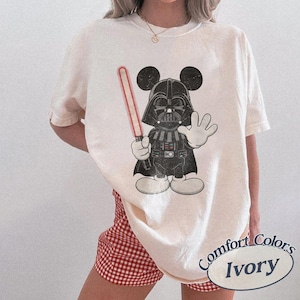 Könnte beinhalten: Ein elfenbeinfarbenes Kurzarm-T-Shirt mit einem Grafikdruck einer schwarzen Darth-Vader-Figur mit Mickey-Maus-Ohren. Die Figur hält ein rotes Lichtschwert in einer Hand und hat die andere Hand erhoben. Das Design hat eine abgenutzte Textur.