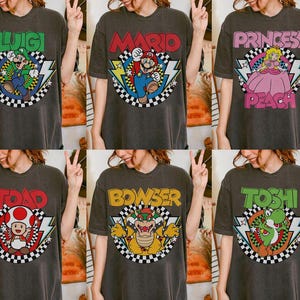May include: A charcoal gray t-shirt featuring a graphic print of a Nintendo character. The design includes the character's name in bold letters, a checkered circle, and lightning bolt accents. Characters include Luigi, Mario, Princess Peach, Toad, Bowser, and Yoshi.