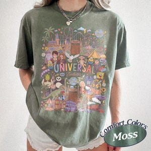 Może przedstawiać: Koszulka z kr&oacute;tkim rękawem w kolorze mchu z kolorowym nadrukiem graficznym przedstawiającym postacie i punkty orientacyjne Universal Studios. Wz&oacute;r zawiera r&oacute;żne animowane postacie, kolejkę g&oacute;rską oraz napis Universal Studios wielokolorową czcionką.