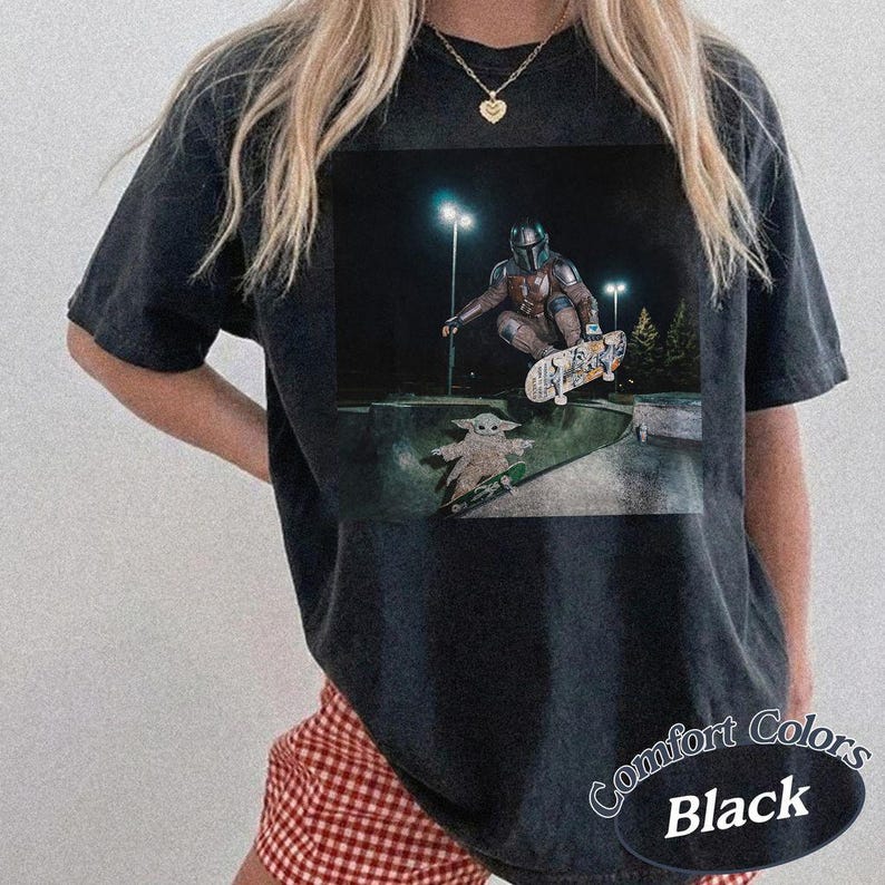 May include: A black t-shirt featuring a graphic print of a character in armor performing a jump on a skateboard in a concrete bowl at night, with a small creature standing on a skateboard below. The shirt has a crew neckline and short sleeves.