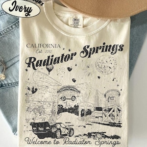 May include: Ivory-colored t-shirt with a graphic of Radiator Springs, California. The design includes the text "Radiator Springs" and "Welcome to Radiator Springs", along with illustrations of cars and buildings. The shirt is made by Comfort Colors.