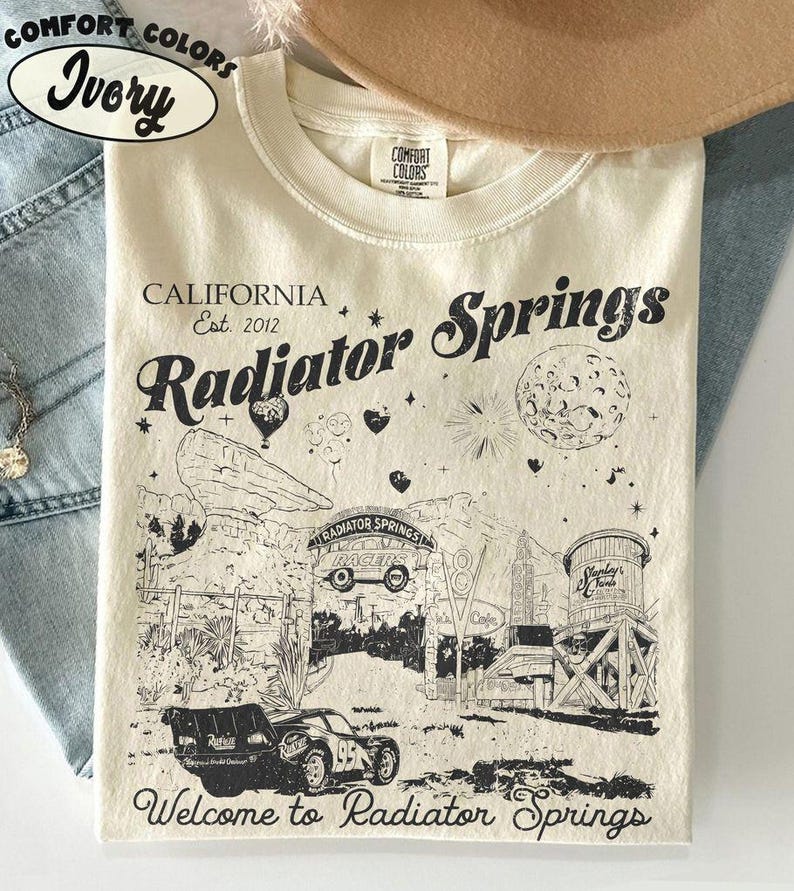 May include: Ivory-colored t-shirt with a graphic of Radiator Springs, California. The design includes the text "Radiator Springs" and "Welcome to Radiator Springs", along with illustrations of cars and buildings. The shirt is made by Comfort Colors.