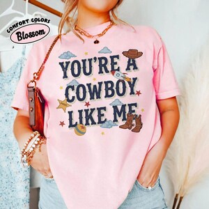 Puede incluir: Una camiseta de manga corta de color rosa claro con el texto azul oscuro "YOU'RE A COWBOY LIKE ME" rodeado de ilustraciones gr&aacute;ficas de un sombrero de vaquero marr&oacute;n, un cohete, una estrella de sheriff amarilla, una bola y botas marrones.