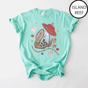 May include: A mint green t-shirt featuring a graphic print of a red cowboy hat, a cow-print cowboy boot with the name Taylor, and a stack of blocks with the letters T, S, and 13. The design includes a beaded string, flowers, and a bow.